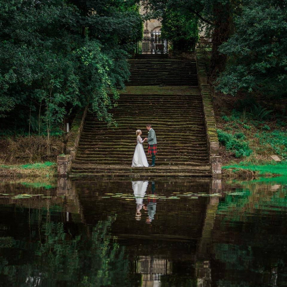 A bride and groom holding hands in at the foot of steps beside a lake